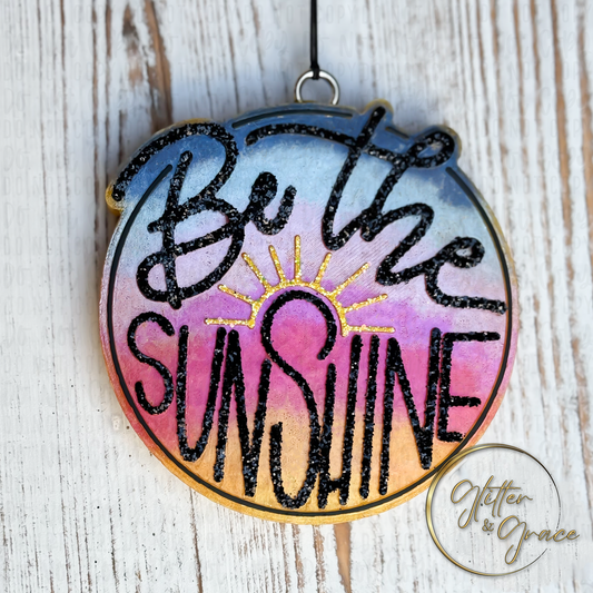 circle shaped vehicle air freshener saying be the sunshine in glitter with the background with sunrise or sunset colors